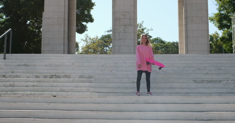 Young blonde long-haired woman dressed in bright pink sweatshirt and trousers dances actively on stairs of park holding penny board
