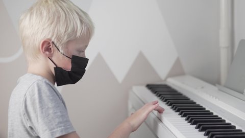 A blonde boy wearing a medical mask on his face plays the piano at school during the covid-19 pandemic.