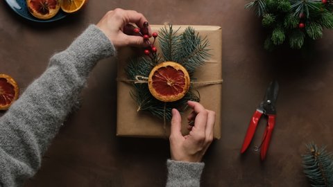 Christmas preparation, packaging of holiday gifts. Female hands decorates Christmas gift box with dried grapefruit and Christmas decoration. Top view, POV. 