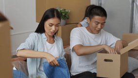 Happy Japanese Family Packing Moving Boxes Sitting Indoor. Couple Preparing For Relocation Putting Clothes In Box, While Children Having Fun In Living Room. Slow Motion