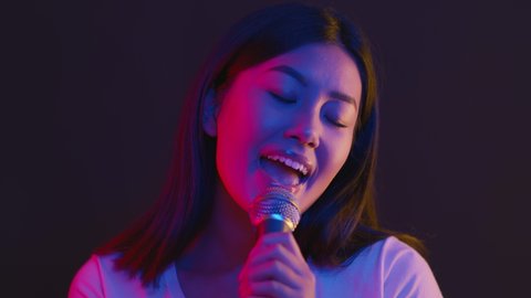 Nightlife entertainment. Young joyful asian woman singing into microphone at karaoke, dancing on stage in bright neon lights, slow motion