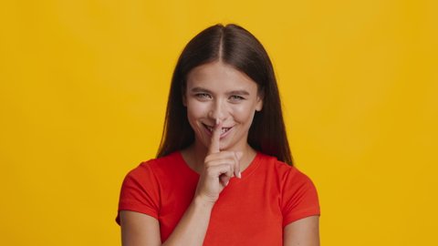 It's A Secret. Lady Gesturing Finger On Lips Smiling To Camera Standing In Studio Over Yellow Background. Headshot Of Smiling Woman Showing Hush Sign Gesture. Keep Silence Concept. Slowmo