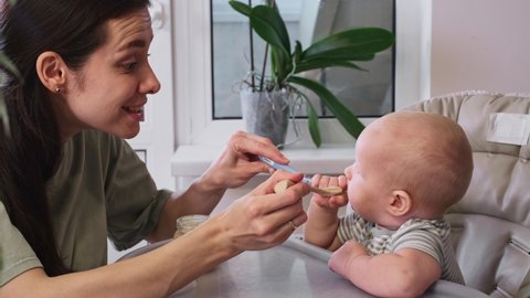 First baby food pure. Mother giving spoon of vegetables or fruits in feeding chair at home. Cute child trying to eat by himself. healthy support. Complementary feeding infant at home. Kids diet