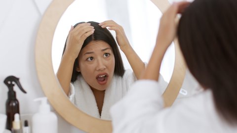 Dandruff problem. Young emotional woman wearing bathrobe looking at mirror and examining her dirty hair with horror, feeling terrified because of scurf, slow motion