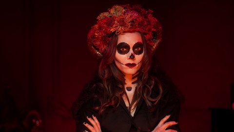 female halloween calavera image in the form of a skeleton with a sewn-up mouth, black eyes, horns, dry flowers on a beautiful Caucasian brunette woman in a dark room with red light