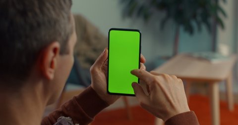 View from shoulder of caucasian man doing swipe up gesture on chroma key of modern smartphone. Mature male resting at home and using cell phone.