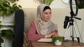 A young Muslim woman in a hijab leads her video blog, live broadcast.Cozy home interior with indoor plants.Slow motion.