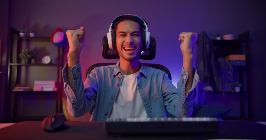 Happy asia guy gamer wear headphone participation play video game colorful neon lights computer in living room at night modern house. Esport streaming game online, Home quarantine activity concept.
