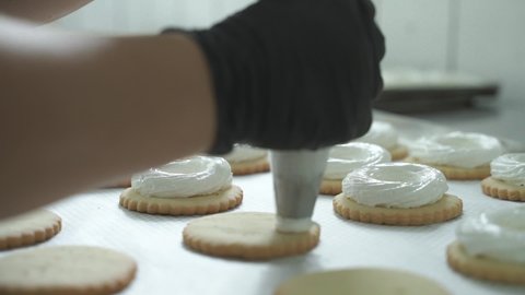Close-up shot of the baker putting cream on top of the cookies with sac a poche. Biscuit production. Confectioner. Delicious cakes and desserts. Pastry chef puts cream on crackers with an icing bag.