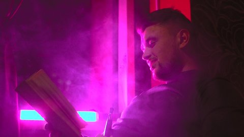Portrait of a young caucasian man reading a book smoking a hookah and blowing out puffs of smoke in the night club with pink lights. Close up shot. Chilling time