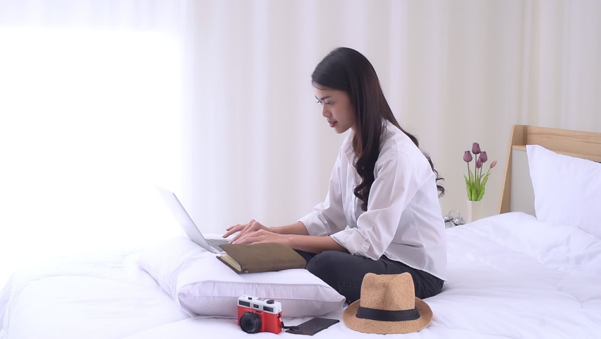 Young Asian woman sitting on the bed and preparing for trip, using a laptop computer to searching information for travel is opening a country after quarantine in hotel during the coronavirus outbreak. Royalty-Free Stock Footage #1081757459