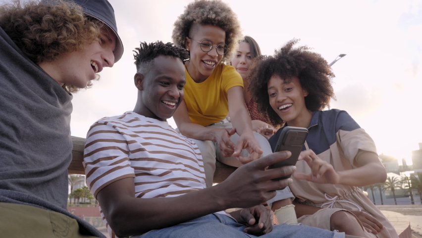 Happy smiling teenage friends laughing outside at something in smartphone or mobile phone. Young multiracial people spending time together. Friendship, communication, youth and lifestyle concept Royalty-Free Stock Footage #1081758380