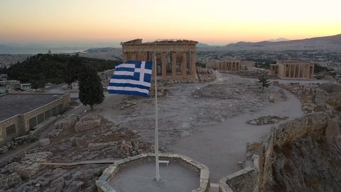 Cinematic shot of the flag of Greece, Acropolis city of Athens parthenon, Mount Lycabettus, Parliament Building and residential buildings, sunset in Athens, Greece. Drone shot
