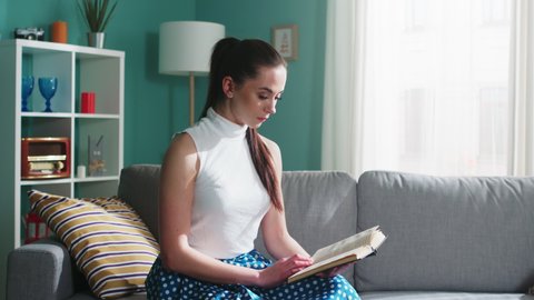 Young intelligent woman in white top is sitting on sofa at home, reading fiction book, deeply captivated by the plot, addicted book-lover, Zoom out, Slow motion.