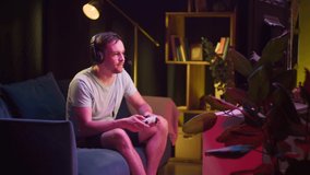 Young man gamer wearing headphones with microphone playing video game on tv, gesturing. Guy using modern console, sitting on sofa in living room. Cybersport, gaming at home concept. 