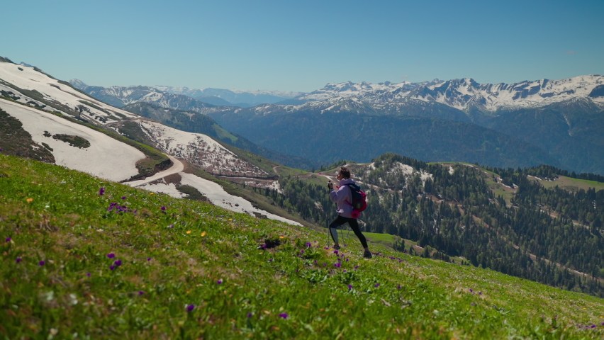 Slow motion shot of a lone girl hiker with trekking poles walking up the green hillside. Mountain journey to enjoy amazing landscapes