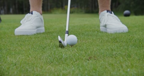 A close-up of a golf club hitting a ball while playing golf. A fair-skinned man in white sneakers plays golf on a green lawn. A dynamic shot of preparing to hit and hit