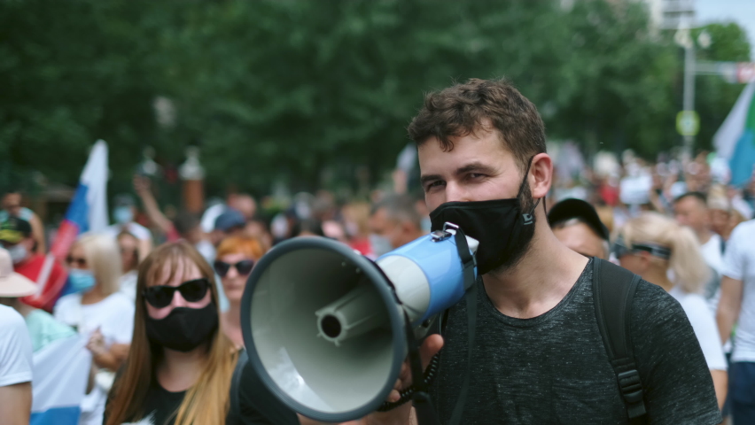 Political strike demonstration rebel guy in Covid-19 restrictions with bullhorn megaphone. Riot demonstrator activist in face mask on opposition resistance protest under lockdown. Rally revolt crowd. Royalty-Free Stock Footage #1081765751