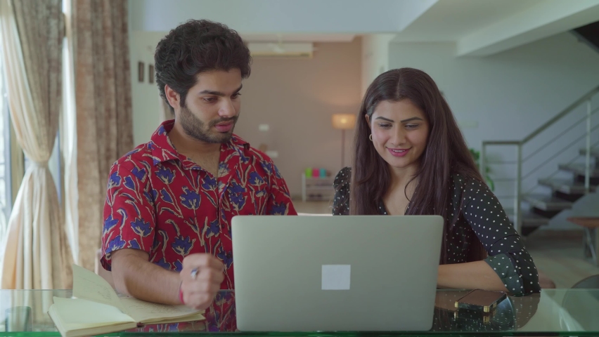 A young modern Asian Indian happy and cheerful couple sitting together in front of a laptop and discussing over an online shopping festival season sale in an interior house