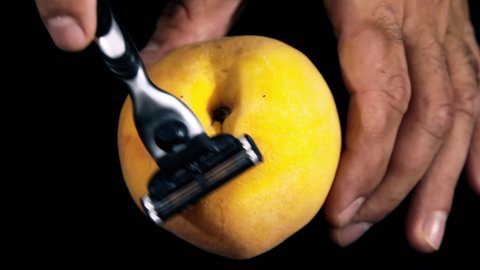 Shaving a peach with a common razor (with the help of hands wearing black gloves). Visual representation (closeup shot) of female or male genitals.

