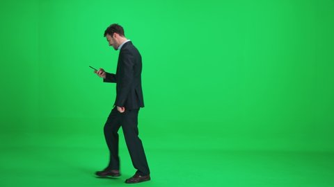 Passer-by businessman in a suit walks on a green background and uses a smartphone, man enjoys financial success and good news, template on a chromakey.