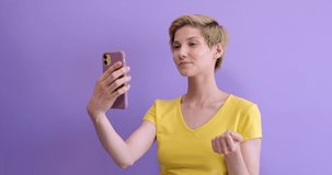 Beautiful caucasian woman in yellow t-shirt having online conversation on phone, video translation with friend or boyfriend, say hello and tell the news. Isolated purple background in studio