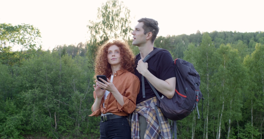 Friendly caucasian couple discussing where to go, using online map on smartphone, handsome man and redhead curly female in casual clothes travelling together, planning the hike. in forest near lake