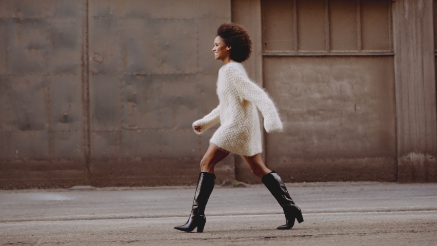 Slow Motion Wide Tracking Shot Of Smiling Young Woman In Woollen Fashion And Boots With Windswept Afro Hair Walking Along Street Royalty-Free Stock Footage #1081773767