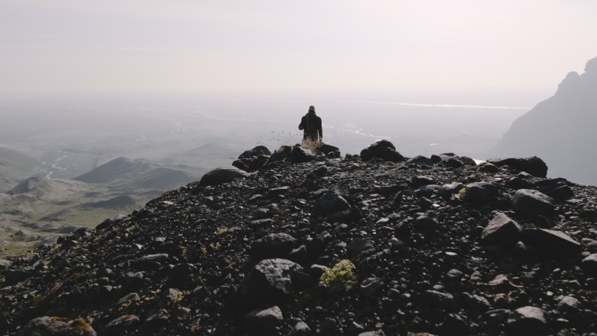 Drone Flying Over Lone Traveller Standing On Rocks With Guitar Case And Looking Down Into Sunlit Valley Of Lakes And Mountains, Iceland