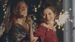 Close up couple of young multiracial girls wearing plush dresses enjoying glamorous Christmas party and having fun. Women dancing with falling confetti and sparkling or glowing sparklers or fireworks. - Powered by Shutterstock - Get 15% off with code: PIKWIZARD15