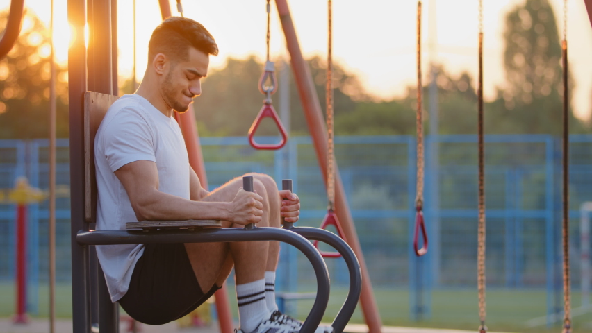 Adult handsome strong sporty Indian guy raises knees high, training abs during morning workout. Middle Eastern athletic young man exercising outdoor at sports ground, pumps up muscles using equipment