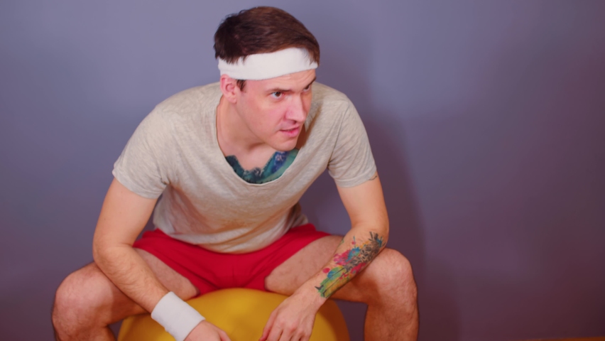 Man in sports headband jumping on gymnastic ball. Active male doing warm-up on fitness ball at home.