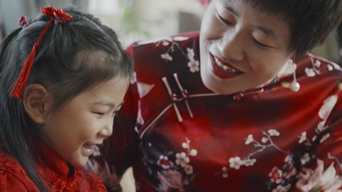 Handheld tilt down shot of happy Chinese mother in qipao dress talking and giving Lunar New Year postcard to her cute 5-year-old daughter in traditional costume