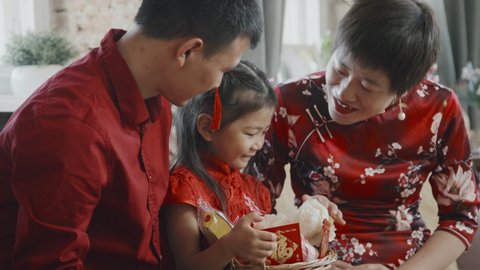 Handheld medium shot of happy Chinese mother and father smiling and chatting with their cute daughter in traditional costume holding gift basket for Lunar New Year