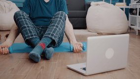 Pensioner looking at training video on laptop to do physical exercise on yoga mat. Old man using dumbbells to lift weights while watching online workout lesson and do pilates activity.