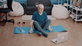 Senior adult using dumbbells to do physical exercise while watching workout video on laptop. Aged man following online training lesson on computer and lifting weights to do fitness activity.