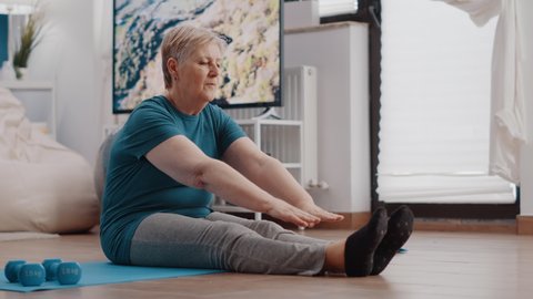 Aged woman doing stretching exercise on yoga mat. Senior person bending over to stretch arms and legs muscles at home for wellness and fitness. Elder adult exercising and training