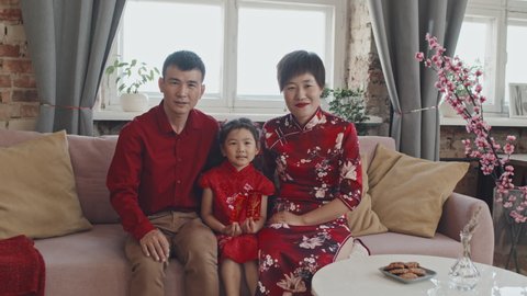 Portrait of Asian parents and 5-year-old girl wearing traditional costumes sitting on couch at home and posing for camera at Chinese New Year celebration