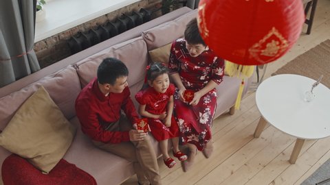Top view shot of happy Asian family of three wearing traditional costumes sitting on couch at home and looking at Chinese New Year postcards