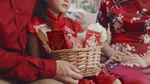 Handheld close up mid-section shot of unrecognizable parents and cute Asian 5-year-old girl in traditional clothing and red shirt sitting together and holding Chinese New Year gift basket