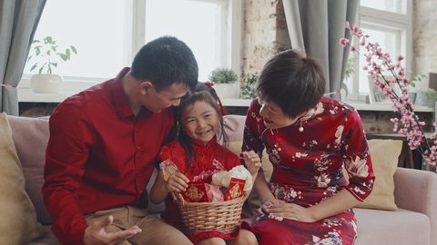 Medium shot of happy Asian father and mother in red shirt and traditional dress giving Chinese New Year gift basket to their cute 5-year-old daughter while celebrating together at home