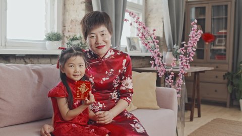 Portrait shot of happy Asian mother and 5-year-old daughter wearing red traditional dresses sitting on couch and posing for camera on Chinese New Year