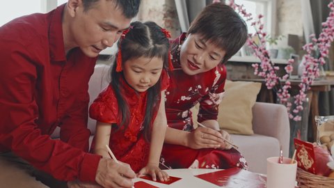 Handheld tilt down medium shot of Chinese parents and cute 5-year-old girl creating Lunar New Year postcard