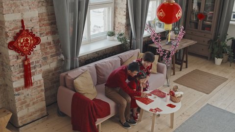 High angle shot of Asian parents and little daughter sitting on couch in living room decorated for Chinese New Year and painting hanzi characters on handmade postcards
