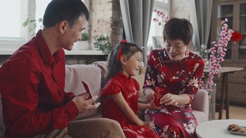 Medium shot with slowmo of happy Asian parents sitting on couch and giving Chinese New Year postcards to cute 5-year-old daughter in traditional costume