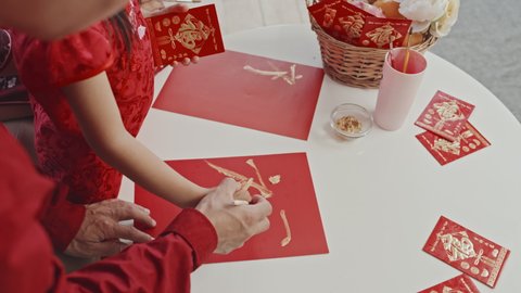 Handheld high angle shot of unrecognizable Asian father helping his little daughter drawing hanzi character on red sheet of paper while making postcard for Chinese New Year