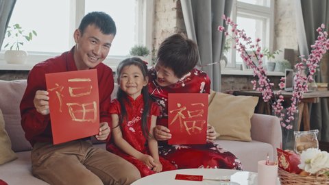 Portrait shot of happy Chinese family with cute 5-year-old girl wearing traditional clothing sitting on couch and showing handmade Lunar New Year postcards to camera