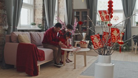 Tracking shot of Chinese family in red clothing sitting on couch in living room decorated for Lunar New Year and making postcards together