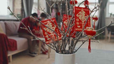 Tracking close up shot with focus on postcards and Chinese New Year decorations hanging on branches in vase. Unrecognizable Asian family with child making postcards in background