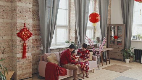 Handheld tracking shot of happy Asian parents in red shirt and traditional dress giving gift basket to cute 5-year-old daughter while celebrating Chinese New Year at home